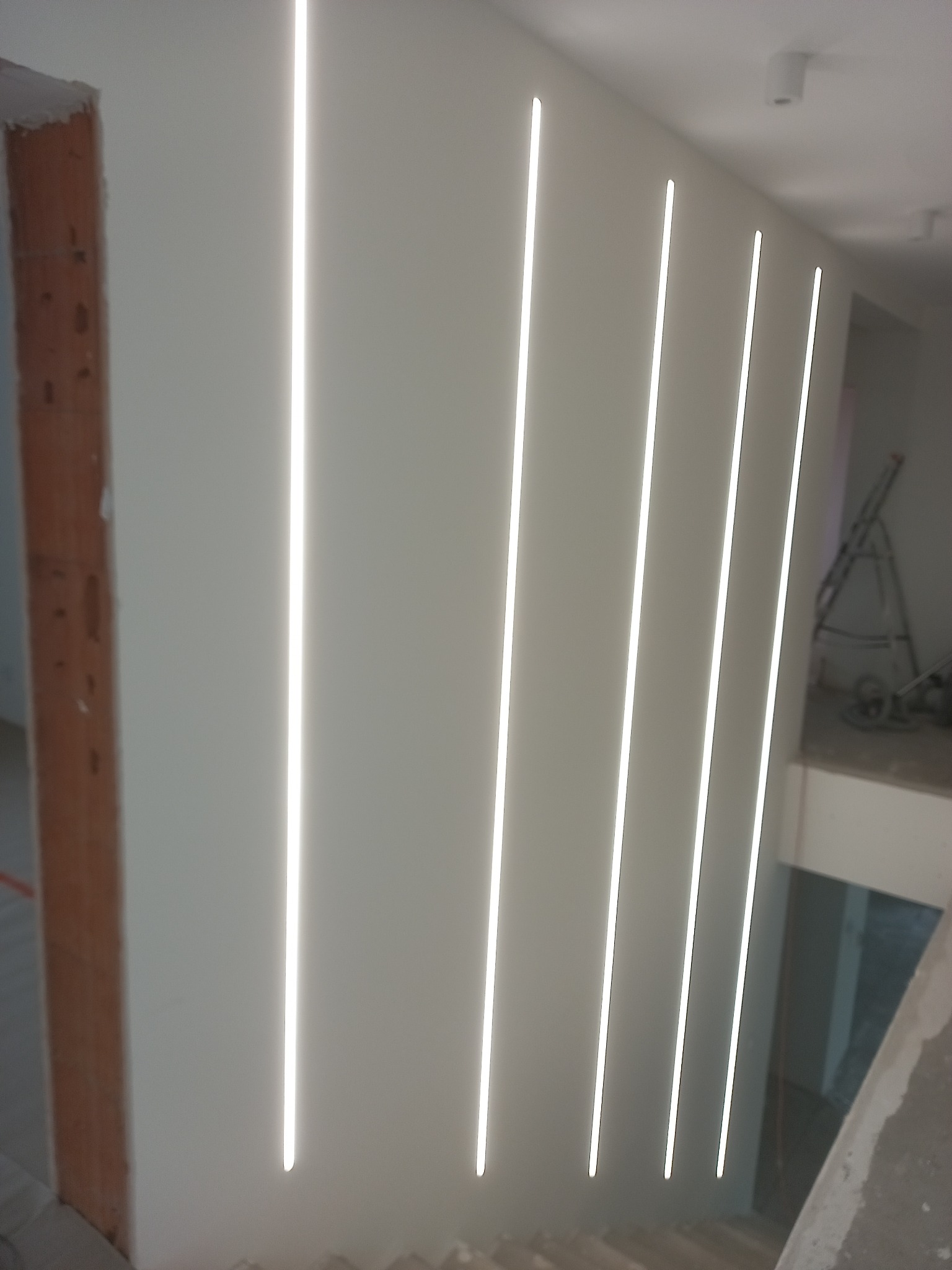 Nowoczesne oświetlenie LED w pionowych liniach na ścianie, widoczne schody i fragment rusztowania w tle. Minimalistyczny design wnętrza w trakcie wykończenia.