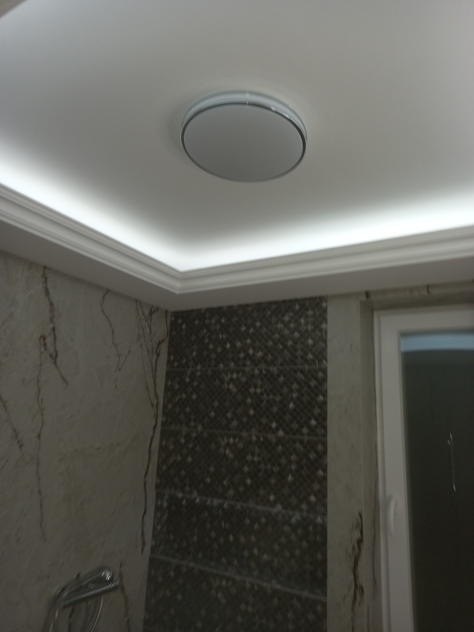 Nowoczesna łazienka z okrągłą lampą sufitową i podświetlanym sufitem. Ściany wykończone marmurem i mozaiką. Widoczny fragment armatury.