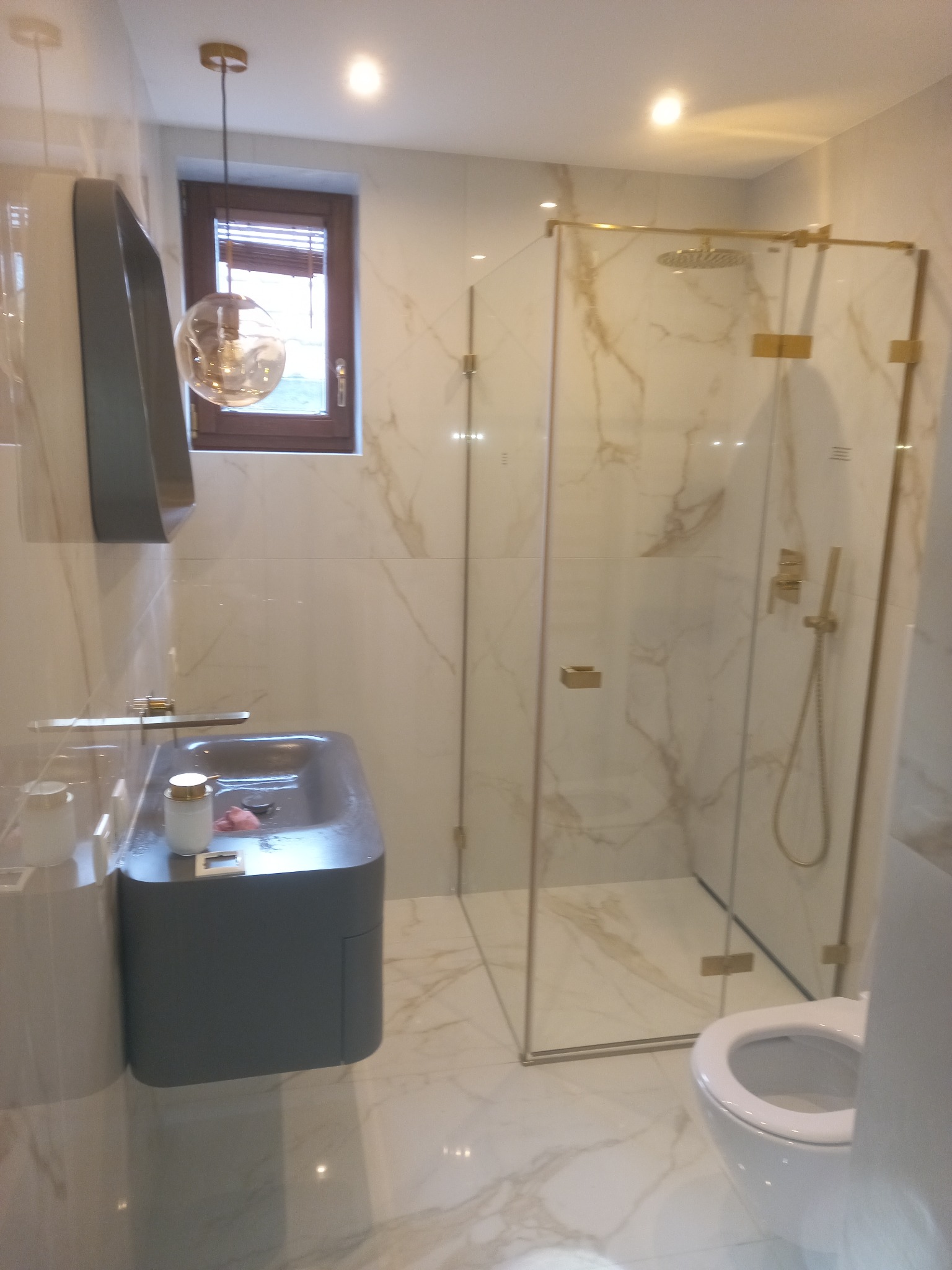 Nowoczesna łazienka z kabiną prysznicową w złotej ramie, umywalką w odcieniach szarości i marmurowymi płytkami. Minimalistyczny design z wiszącą lampą.