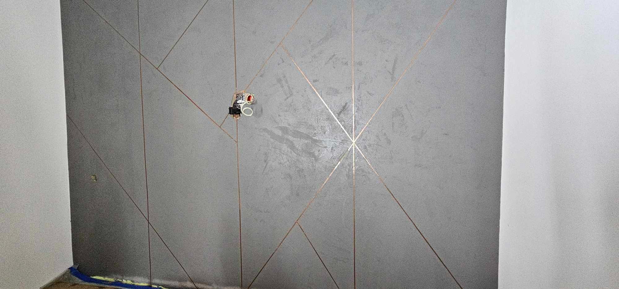 Szara ściana z dekoracyjnymi, złotymi liniami tworzącymi geometryczny wzór. Widoczne gniazdko elektryczne i fragment podłogi z niebieską taśmą malarską.