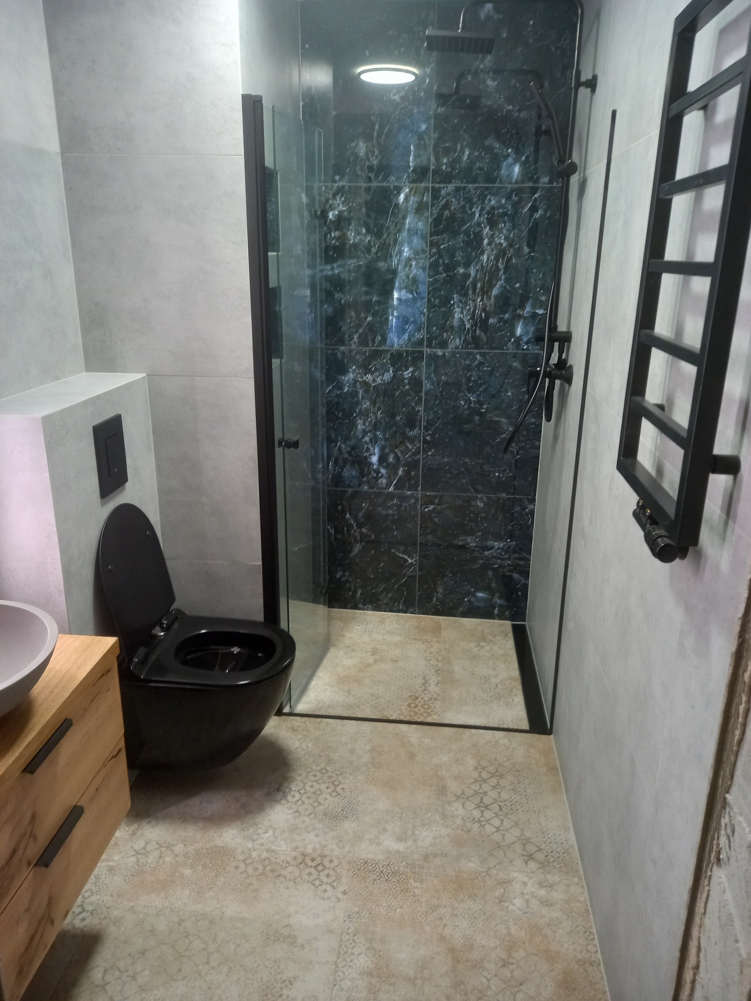 Nowoczesna łazienka z czarną armaturą: toaleta, grzejnik drabinkowy i kabina prysznicowa z marmurowymi płytkami. Jasne płytki podłogowe z delikatnym wzorem.