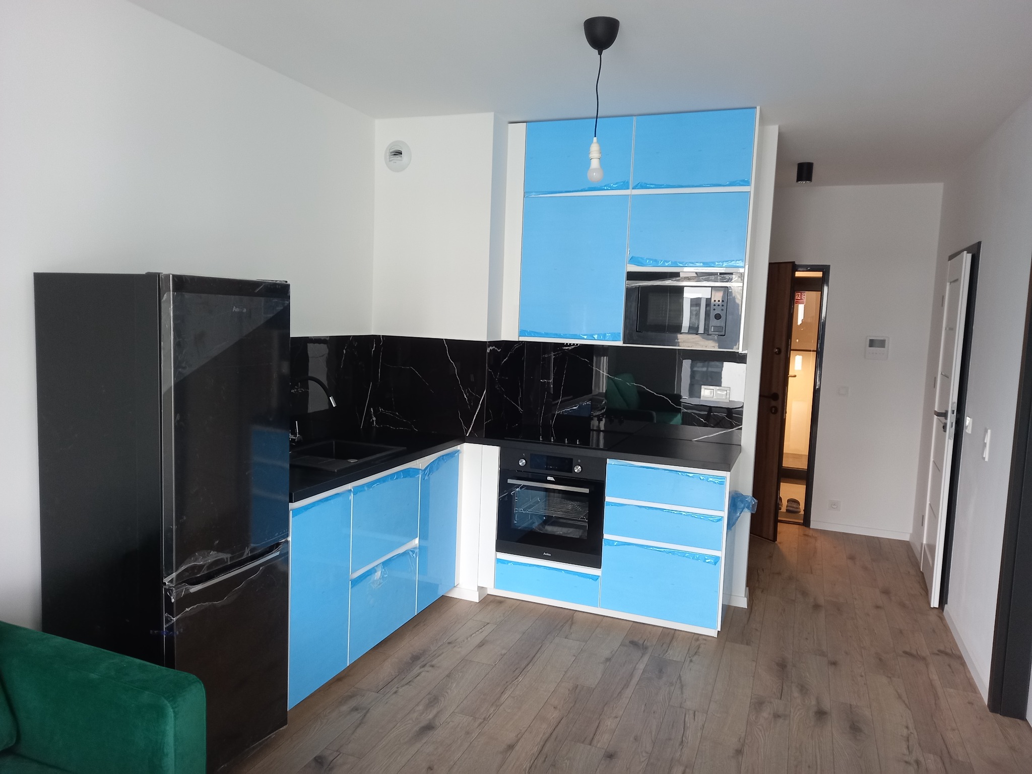 Nowoczesna kuchnia z czarnym blatem i sprzętami AGD, zabezpieczona folią ochronną. Widoczne drewniane panele podłogowe i minimalistyczne oświetlenie.