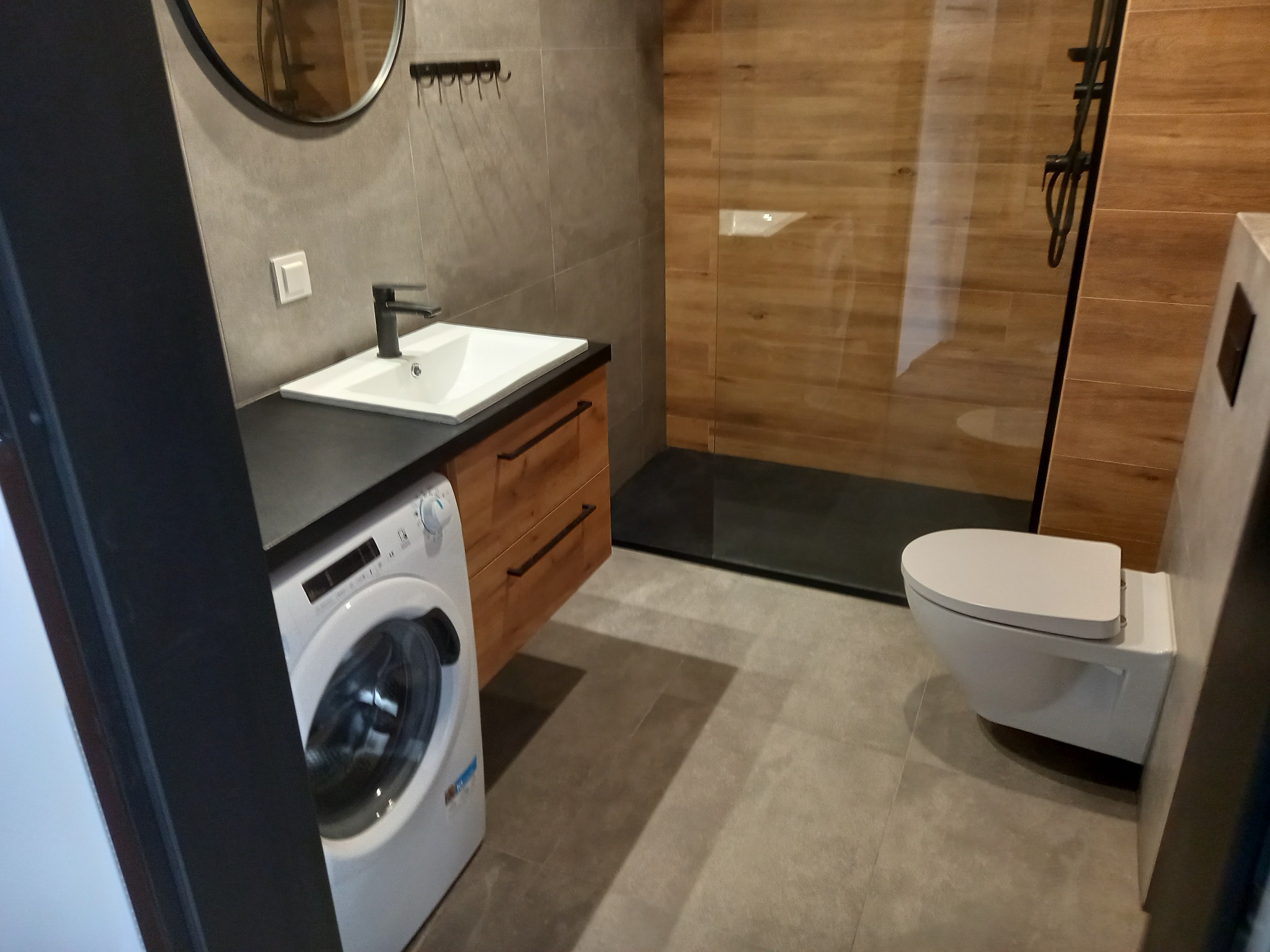 Nowoczesna łazienka z drewnianymi akcentami: kabina prysznicowa typu walk-in, podwieszana toaleta, pralka i umywalka z szafką. Szare płytki na ścianach i podłodze.