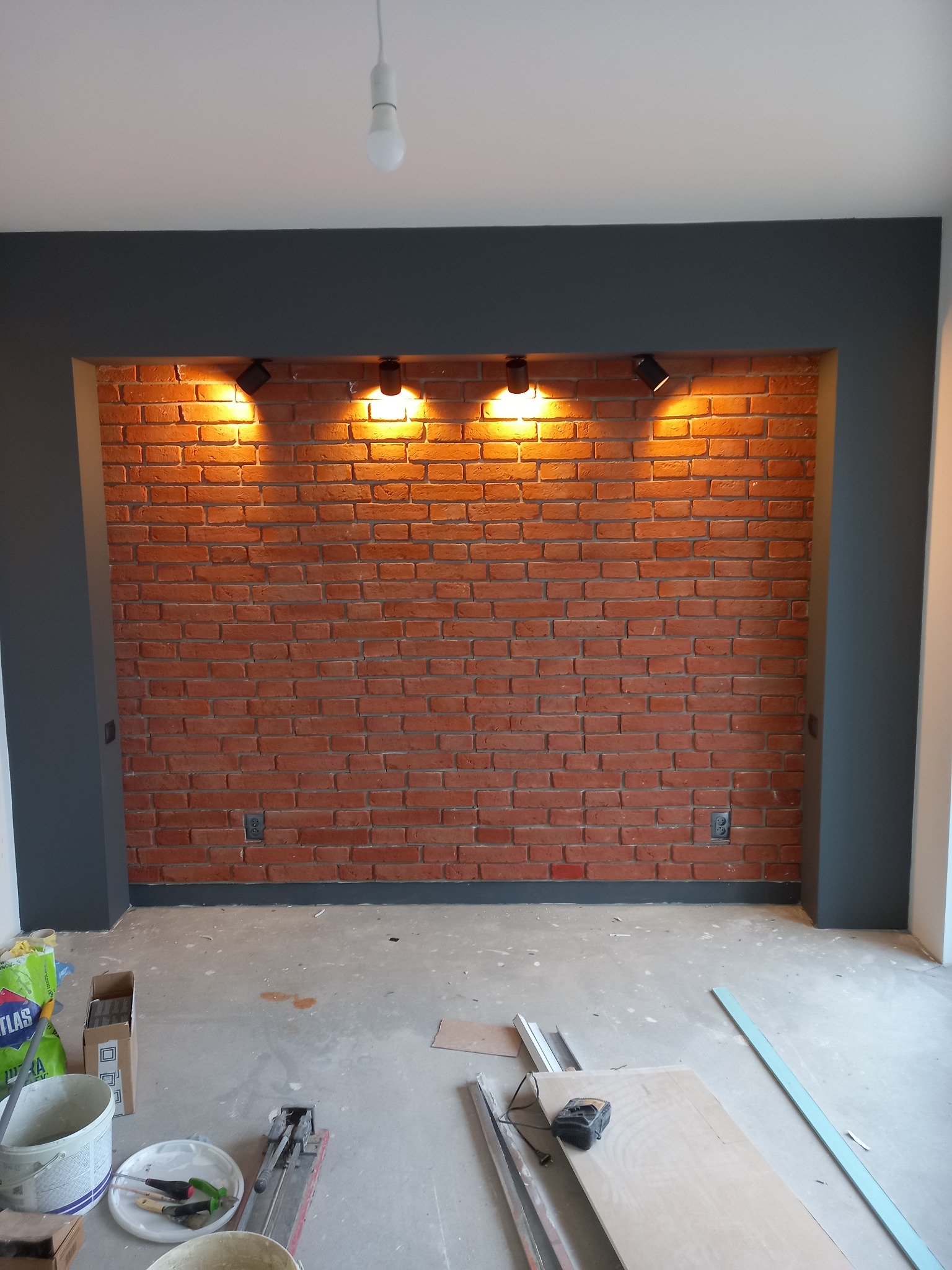 Ceglana ściana z oświetleniem punktowym w ramie pomalowanej na ciemny kolor. Na podłodze widoczne narzędzia i materiały budowlane.