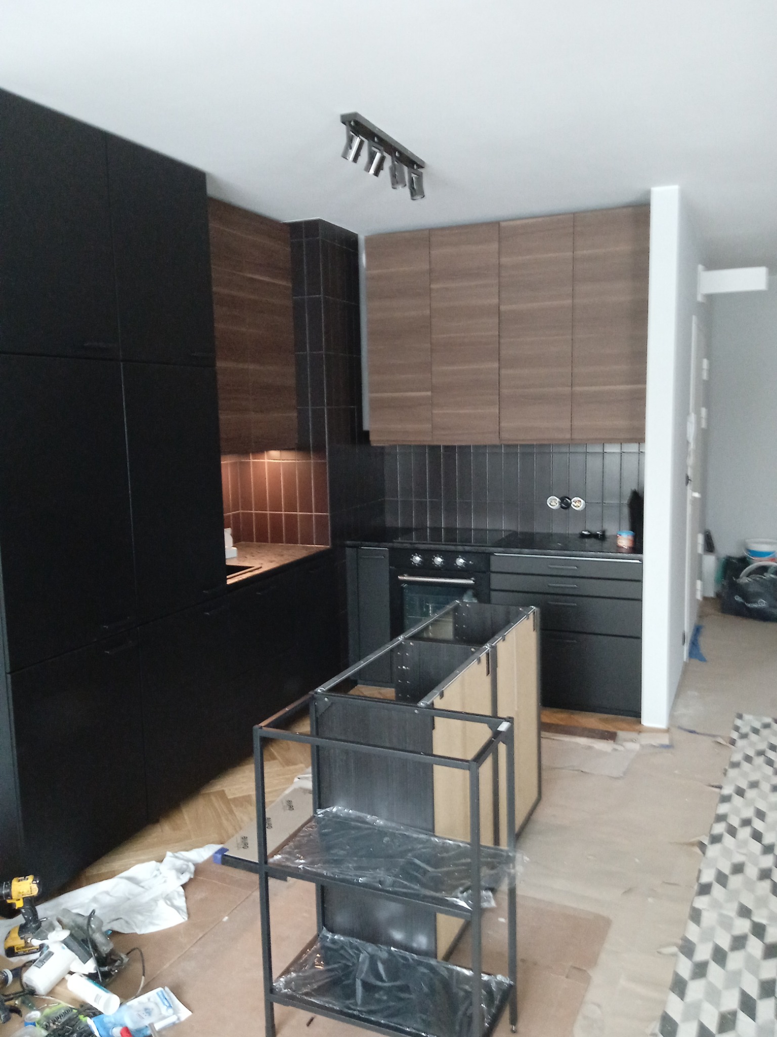 Częściowo zmontowana wyspa kuchenna na tle nowoczesnej zabudowy z ciemnymi szafkami i brązowymi panelami. Widoczne narzędzia i materiały montażowe.