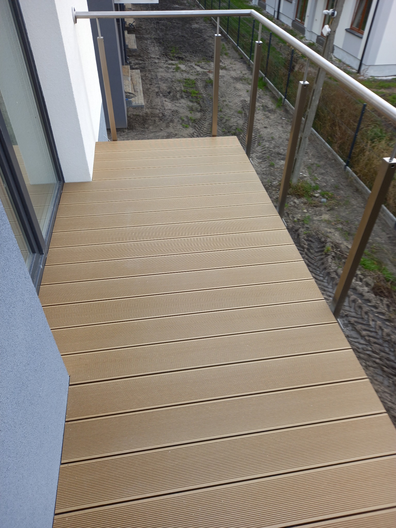 Nowoczesny balkon z desek kompozytowych w odcieniach beżu, wykończony metalową balustradą. Widok z góry na ułożone deski i fragment elewacji budynku.