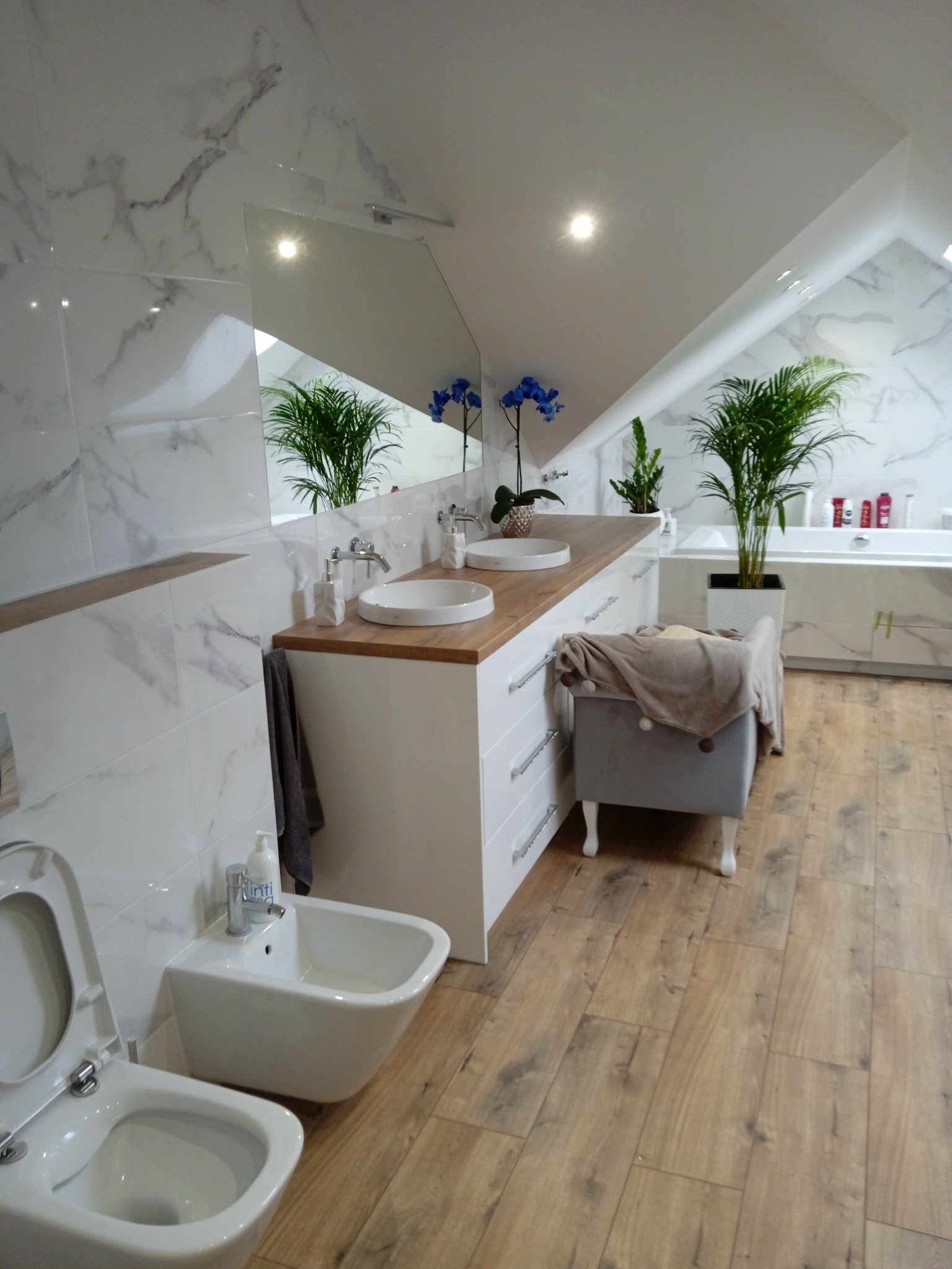 Jasna łazienka na poddaszu z drewnianą podłogą, marmurowymi płytkami i białymi meblami. Umywalki nablatowe, bidet i toaleta w nowoczesnym stylu. Rośliny doniczkowe dodają świeżości.