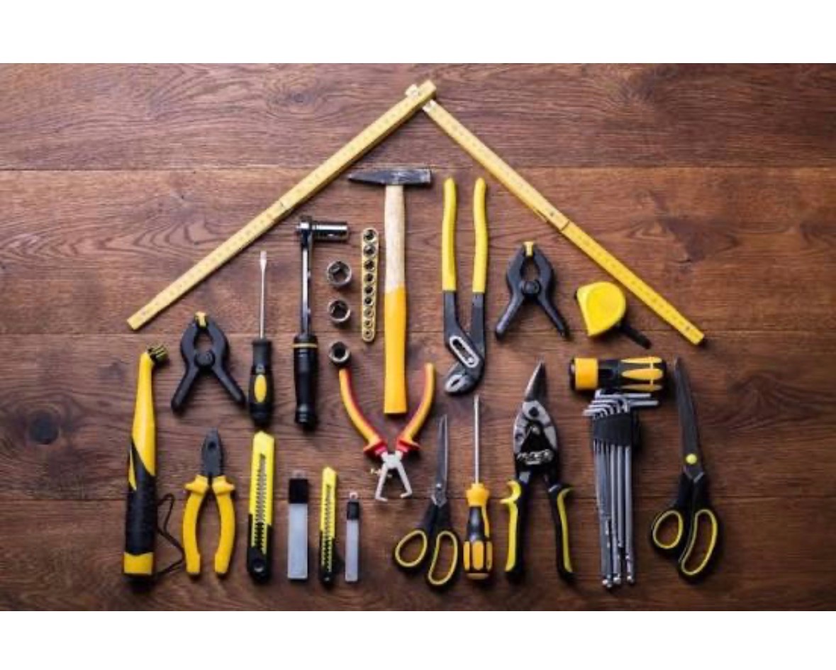 Zestaw narzędzi ręcznych z żółtymi akcentami, ułożonych na drewnianym blacie w kształt domu. Widoczne: młotek, śrubokręty, kombinerki, miara składana i inne.