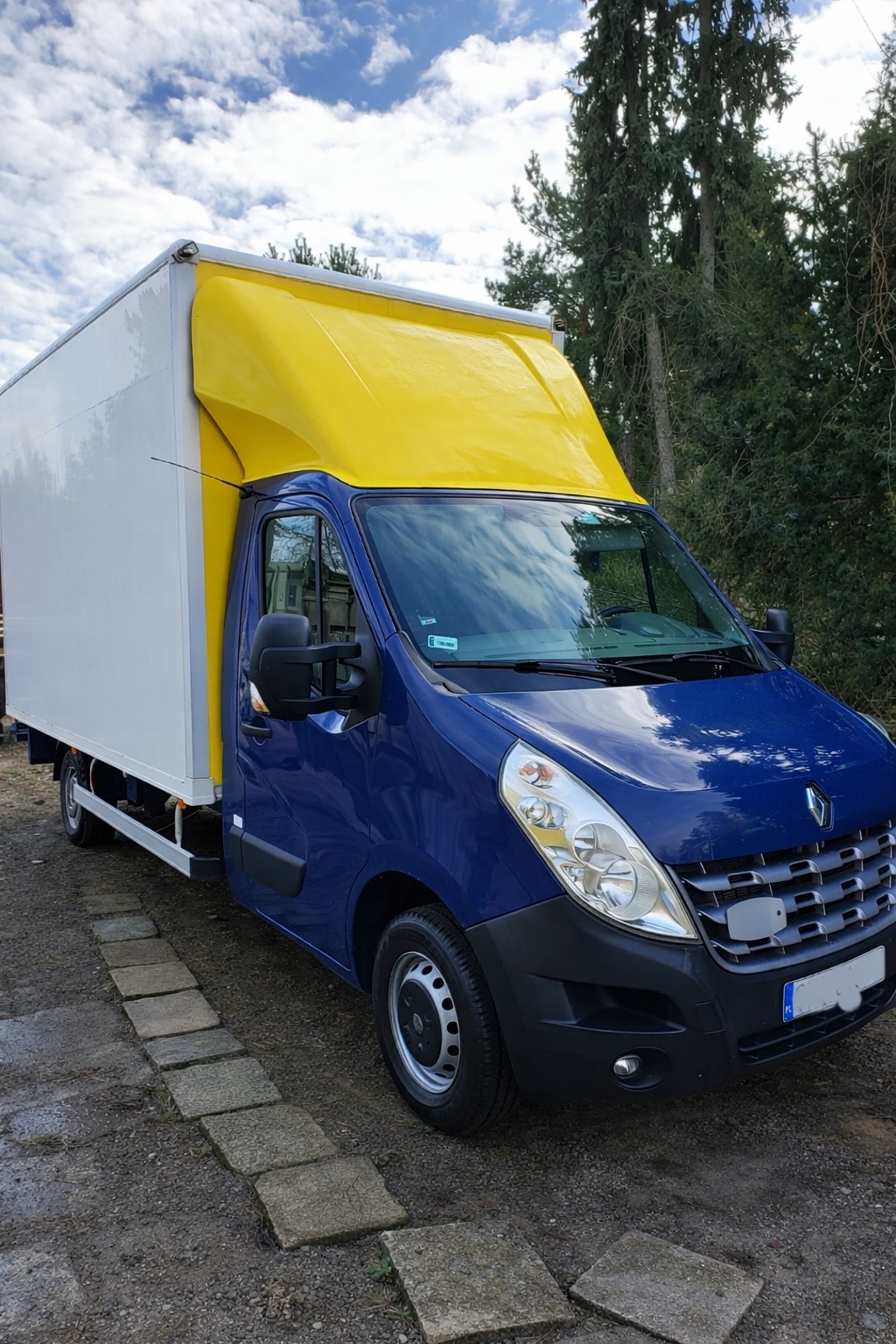 Niebieski bus Renault Master z białą skrzynią i żółtą plandeką na tle drzew i błękitnego nieba z chmurami, zaparkowany na szarym podłożu z kamiennymi płytami.