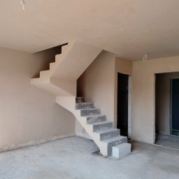 Arkadiusz - Wnętrze po nałożeniu gładzi gipsowej: ściany, sufit i schody pokryte jasnym tynkiem. Widoczne niedoskonałości przy podłodze i ościeżnicach. Surowy stan wykończenia.