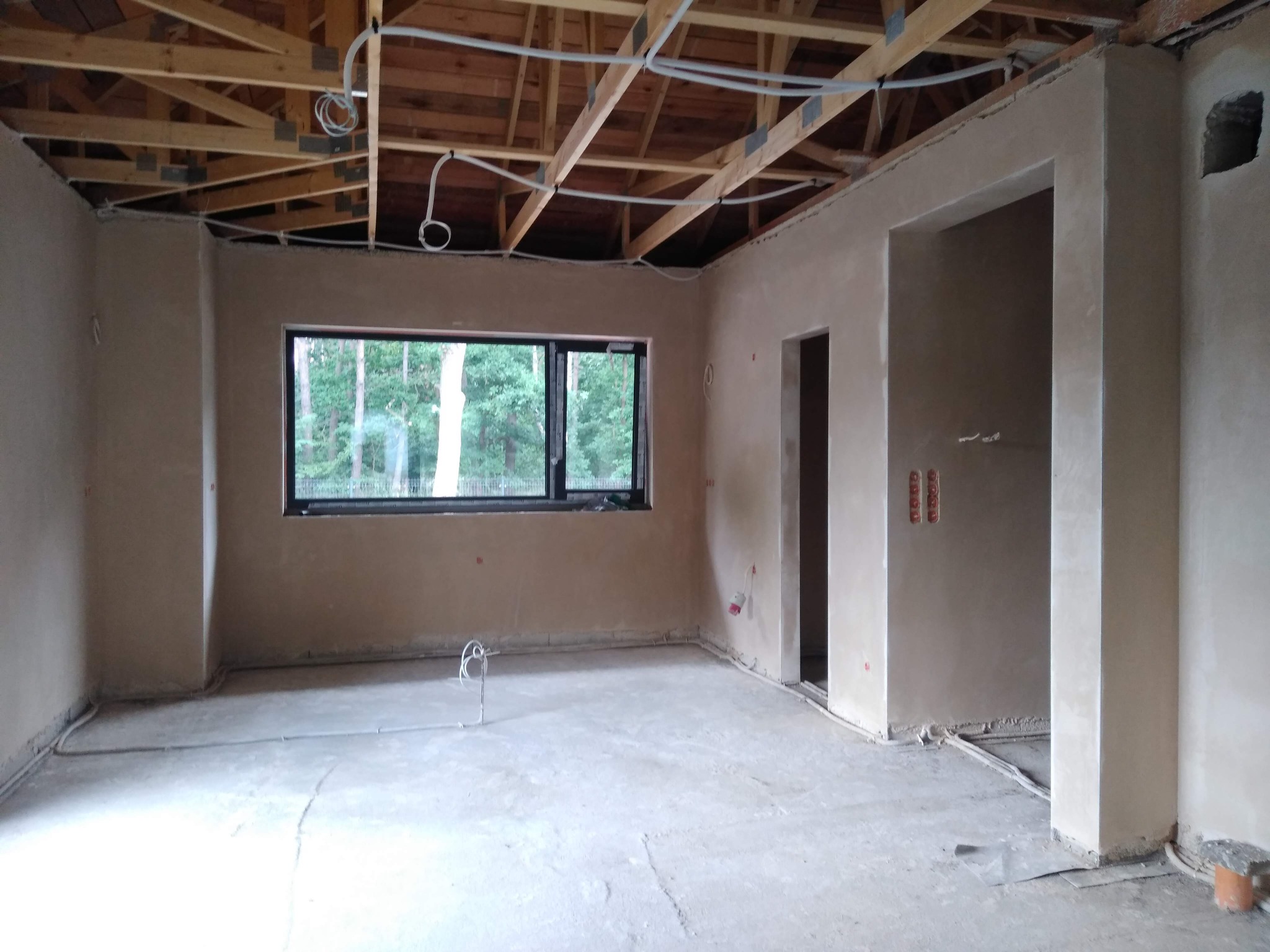 Wnętrze w trakcie prac wykończeniowych: ściany pokryte świeżym tynkiem, widoczne instalacje elektryczne pod sufitem i przy podłodze, surowy stan podłogi.