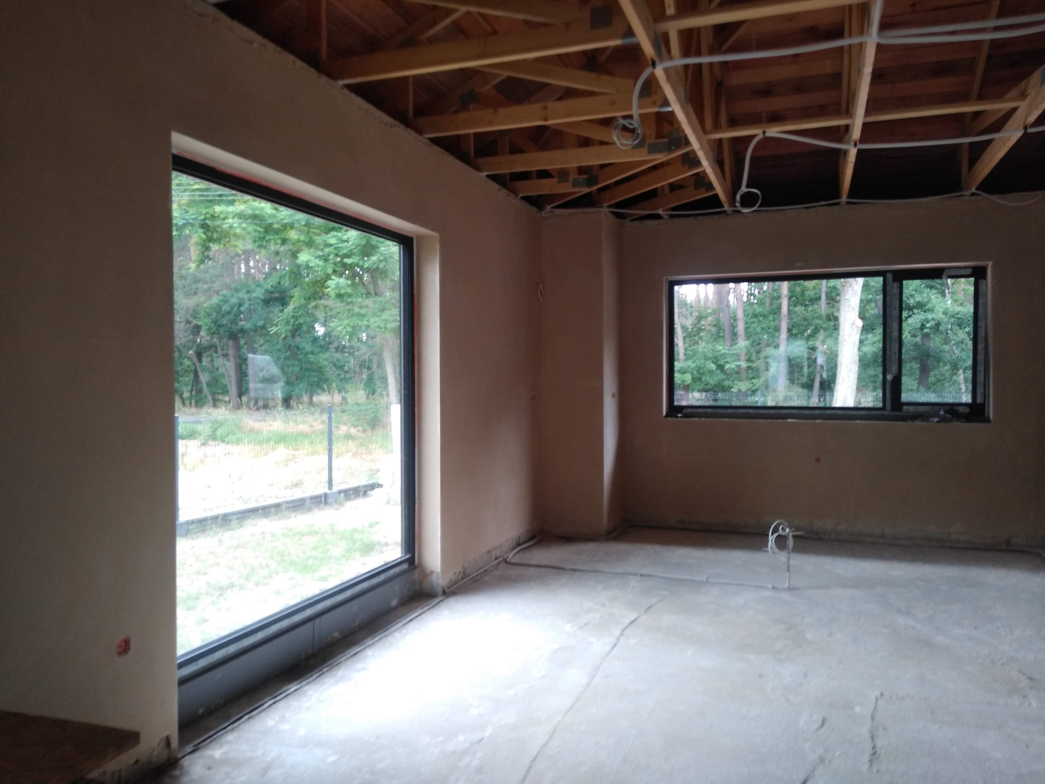 Surowe wnętrze z oknami i drewnianą konstrukcją dachu po nałożeniu tynków. Widoczne instalacje elektryczne. Tło stanowią drzewa za oknami.