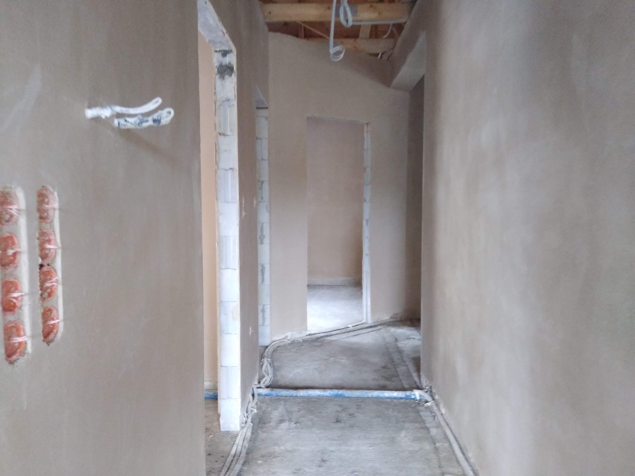 Wnętrze w trakcie prac wykończeniowych: świeżo położone jasne tynki na ścianach, widoczne instalacje elektryczne i rury na podłodze, surowy stan wykończenia.