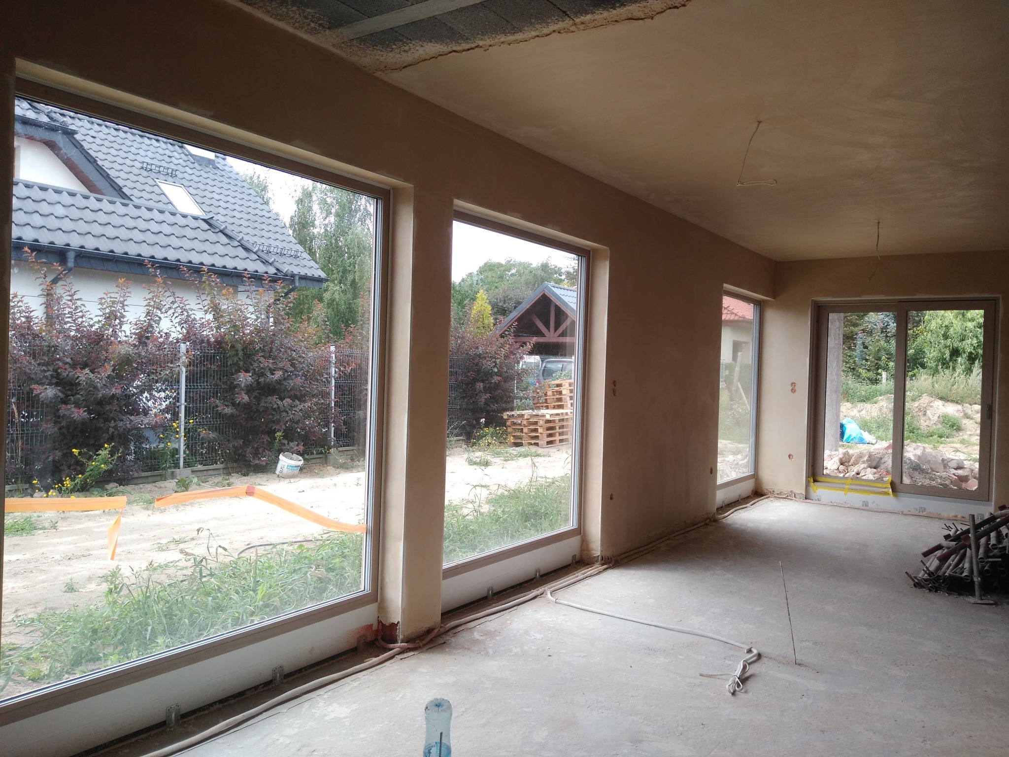 Wnętrze domu po nałożeniu tynków, widoczne duże okna z widokiem na zewnątrz. Ściany i sufit pokryte świeżym tynkiem, podłoga surowa, widoczne instalacje.