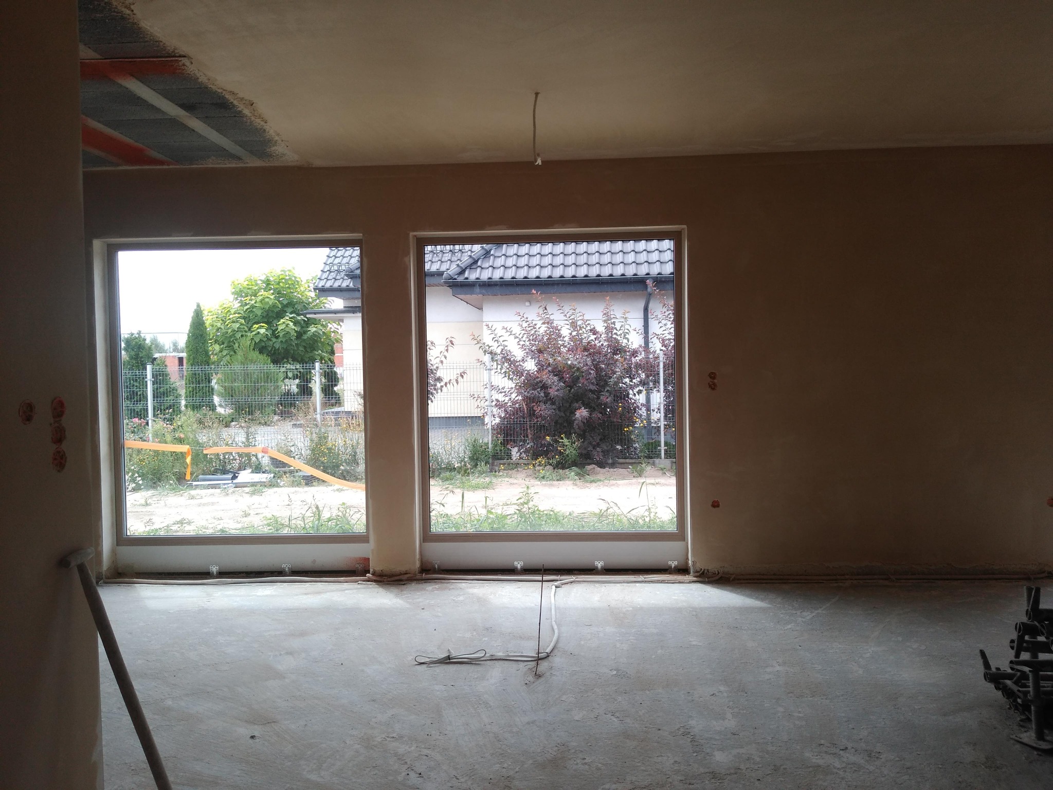 Wnętrze po nałożeniu tynków: gładkie ściany, widoczne okna z widokiem na zieleń, surowy beton na podłodze, wystające przewody elektryczne.