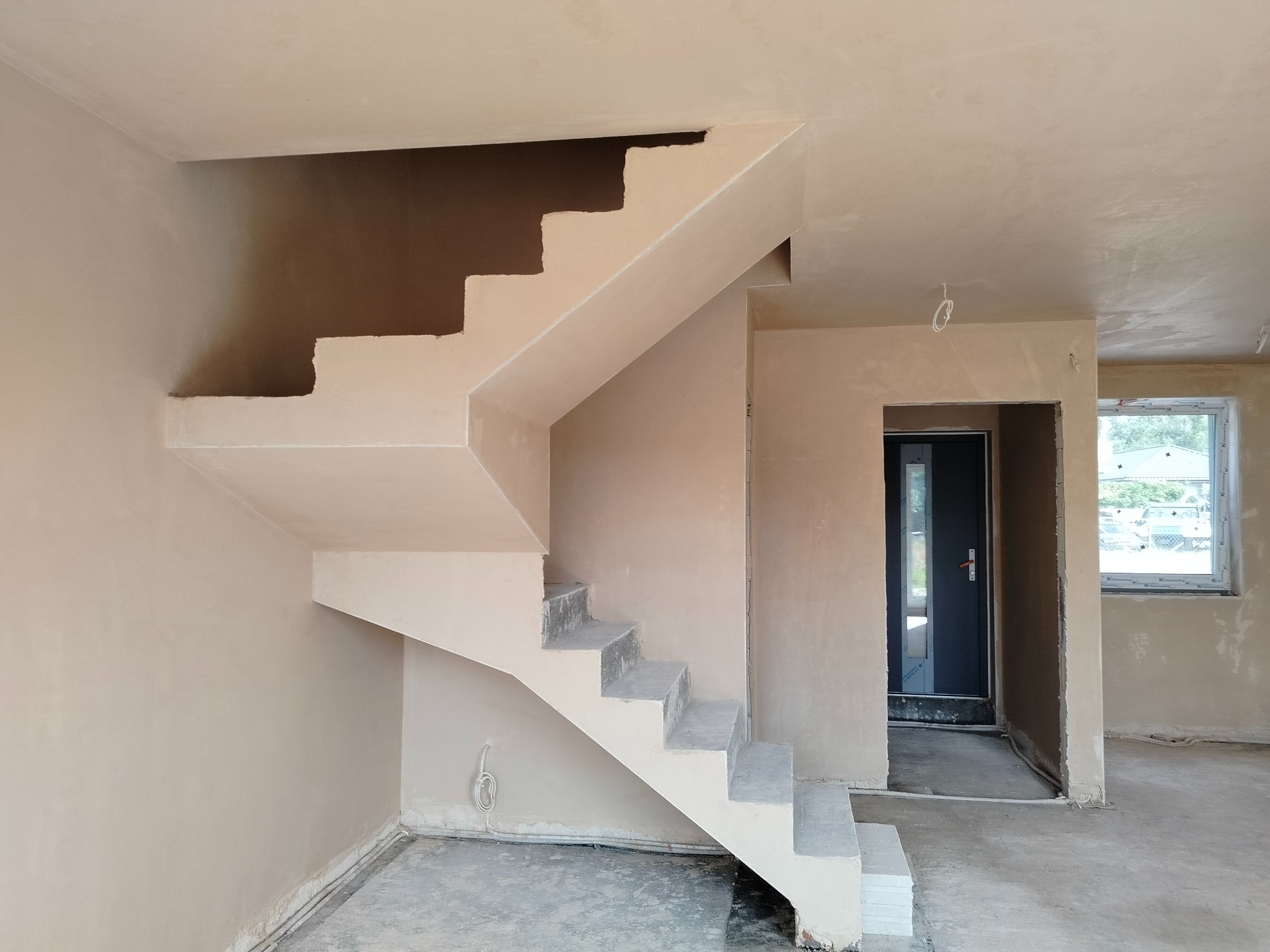 Wnętrze po nałożeniu jasnych tynków na ściany i schody. Widoczne surowe schody przed wykończeniem, drzwi wejściowe i okno. Ściany gładkie, przygotowane do dalszych prac.