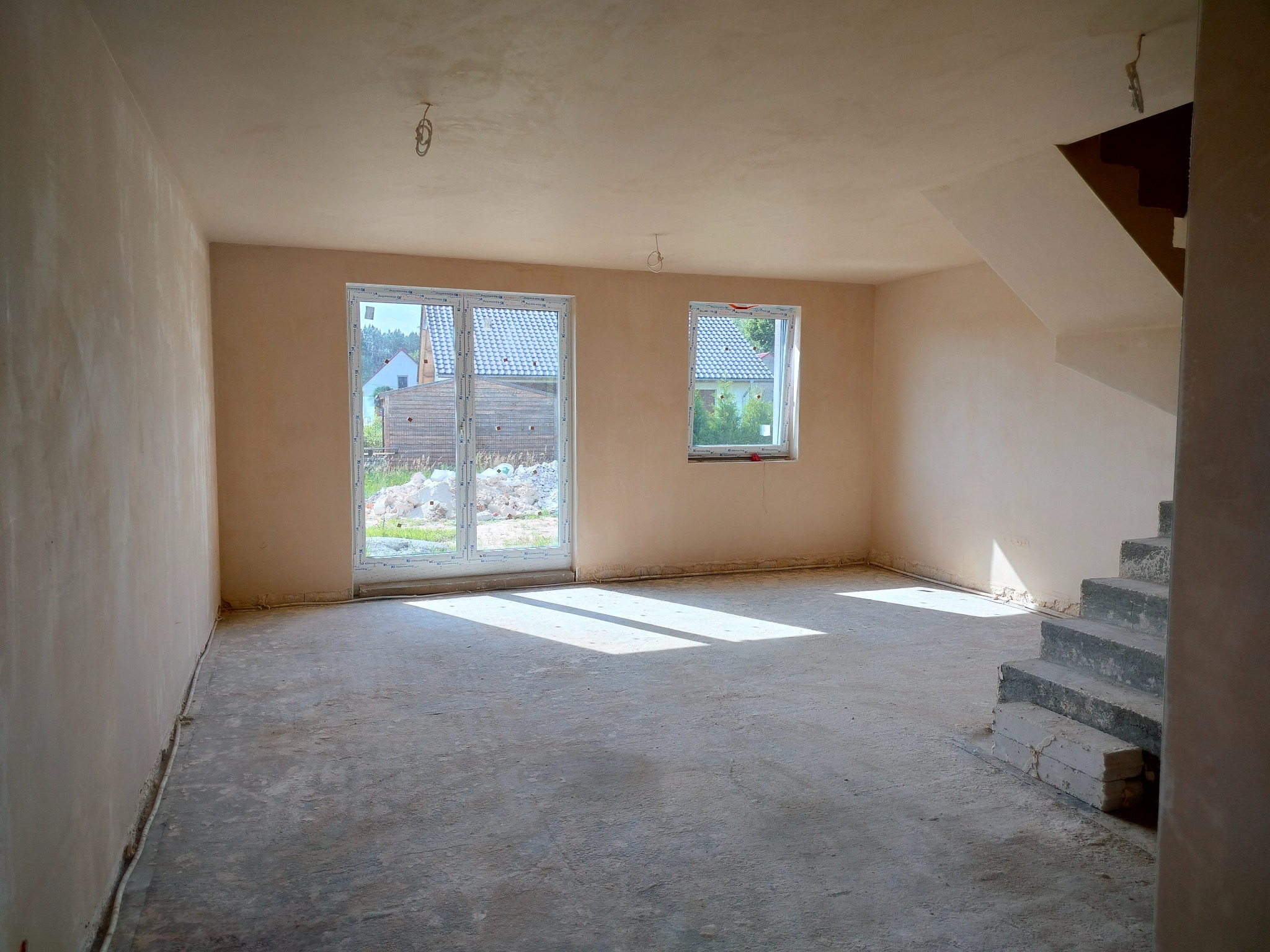 Wnętrze po nałożeniu tynków: jasne ściany, okna z widokiem na zewnątrz, surowy beton na podłodze, schody w stanie surowym. Widoczne niedoskonałości i detale wykończenia.