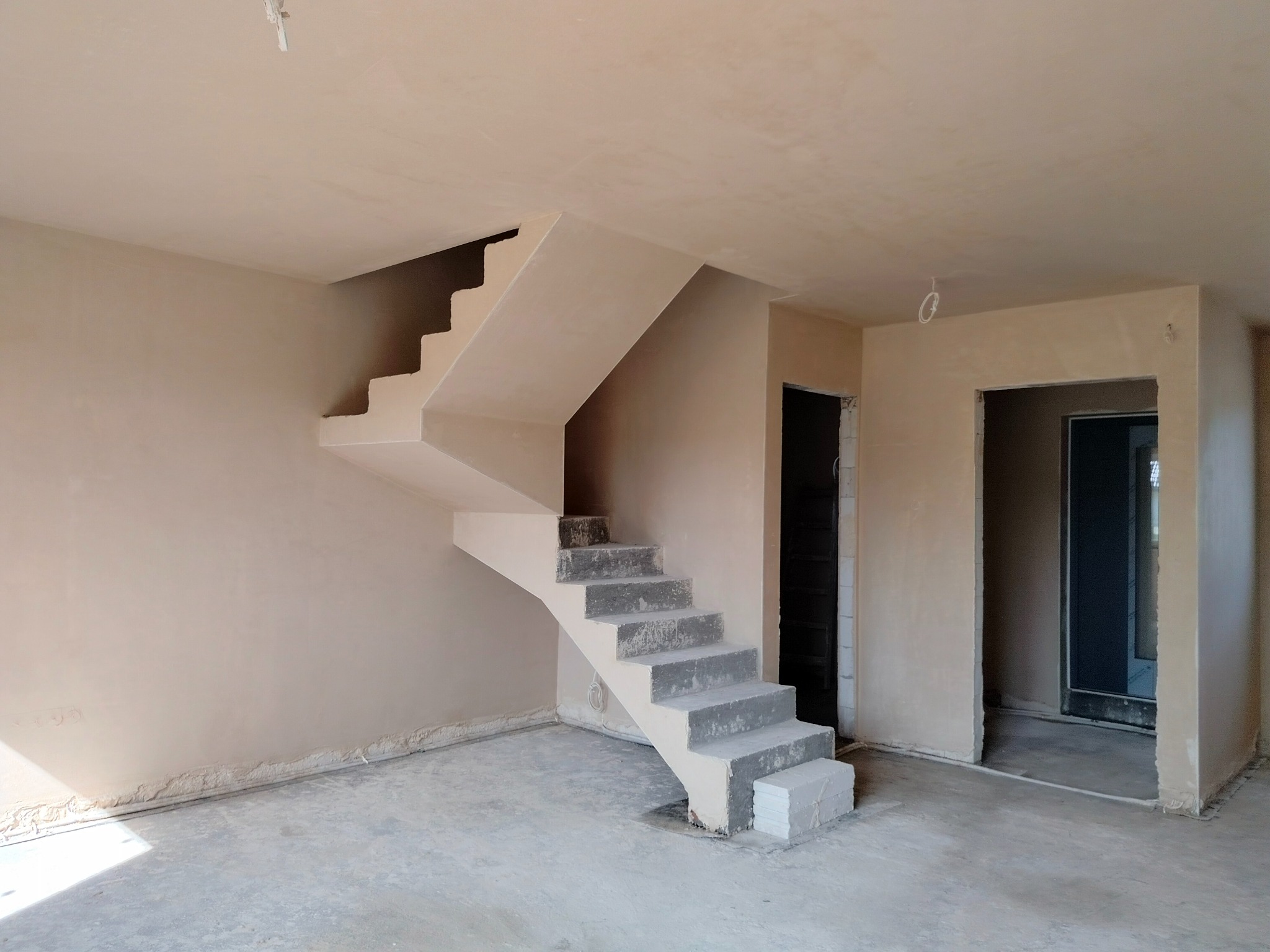 Wnętrze po nałożeniu gładzi gipsowej: ściany, sufit i schody pokryte jasnym tynkiem. Widoczne niedoskonałości przy podłodze i ościeżnicach. Surowy stan wykończenia.