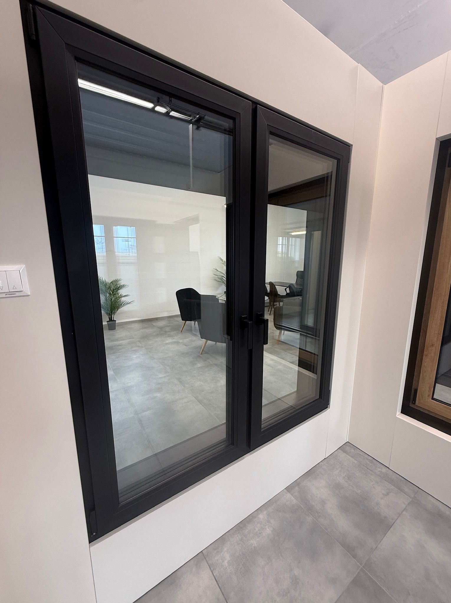 Nowoczesne okno PCV z czarną ramą, widok z boku, prezentacja w salonie. W tle widoczne wnętrze biura z szarymi meblami i roślinami doniczkowymi.