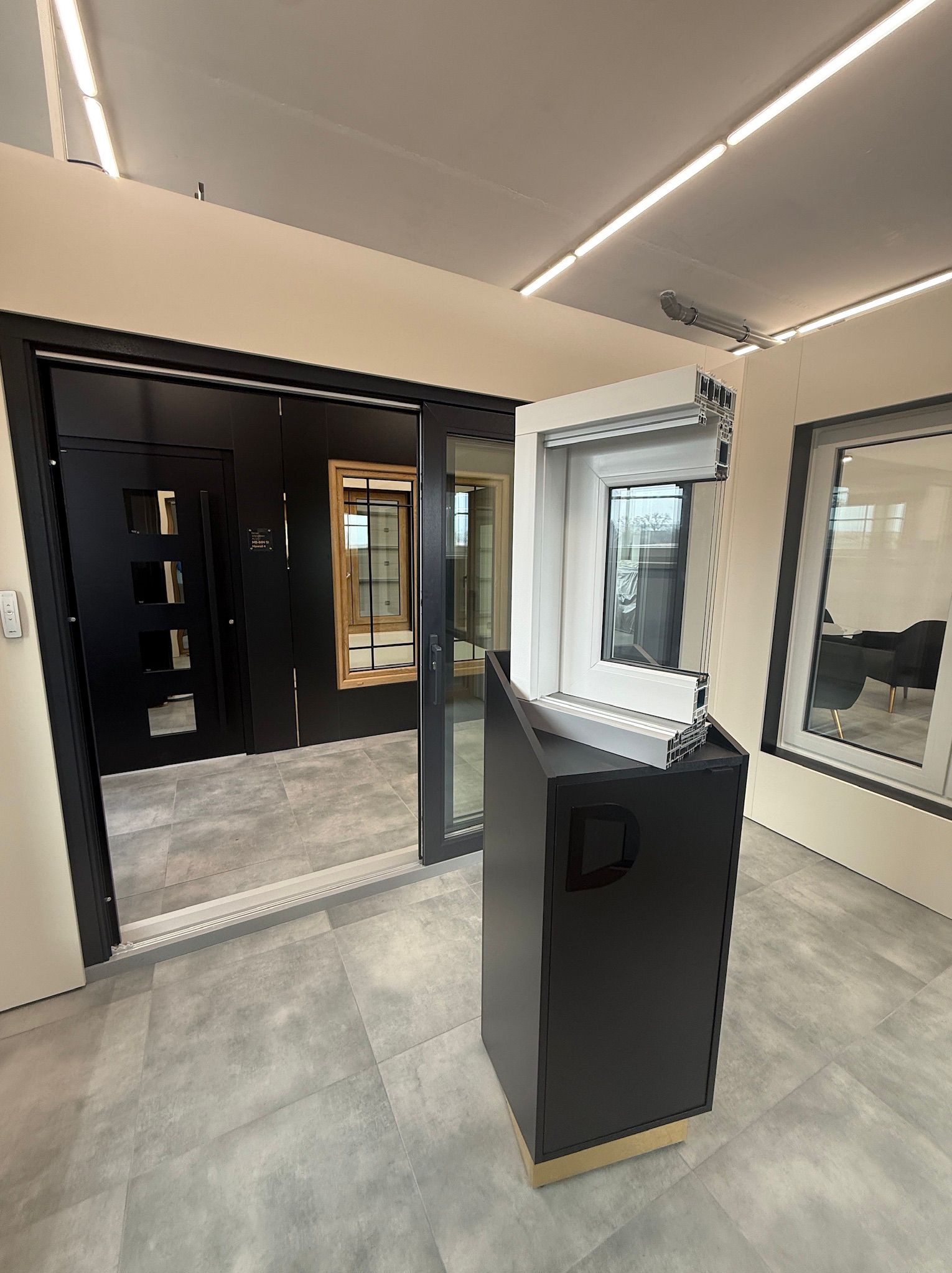 Ekspozycja przekroju okna PCV na czarnym postumencie w salonie, obok drzwi przesuwnych i okna. Widoczna struktura profilu okiennego i detale wykończenia.