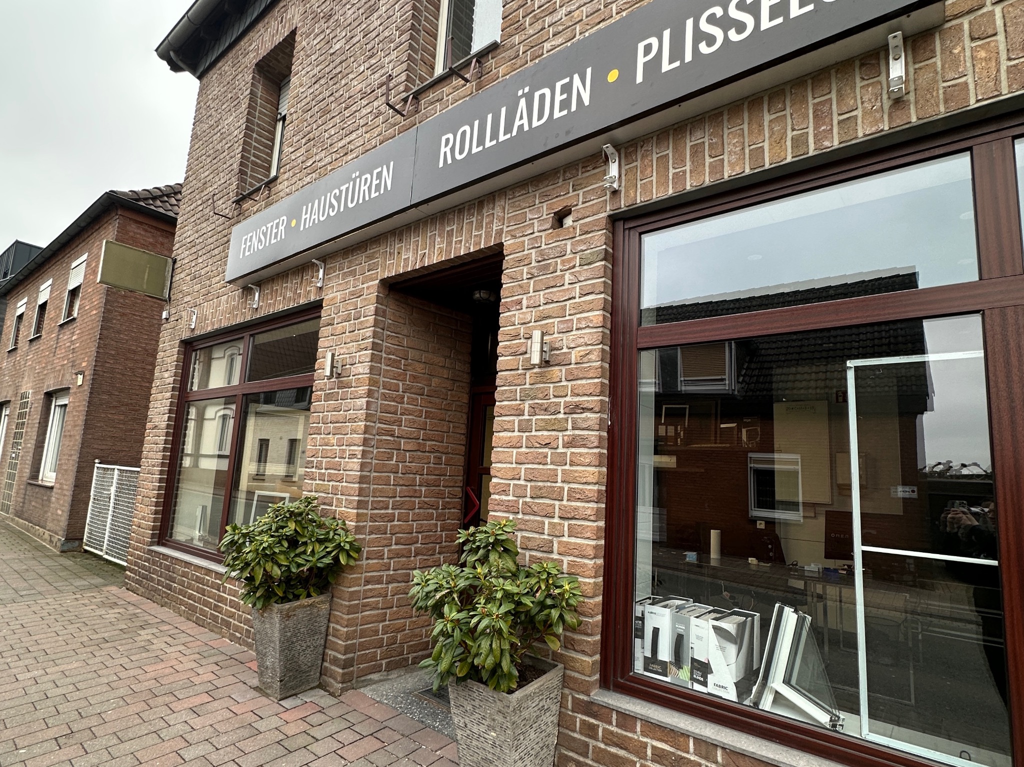 Fasada budynku z cegły z oknami i drzwiami. Nad wejściem szyld z napisem 'ROLLÄDEN • PLISSEES' i 'FENSTER • HAUSTÜREN'.