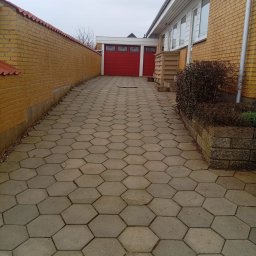 Marian Moskwa Contractor - Ułożona szara kostka brukowa w kształcie plastra miodu na podjeździe. W tle czerwona brama garażowa i ceglany mur. Widok z perspektywy.
