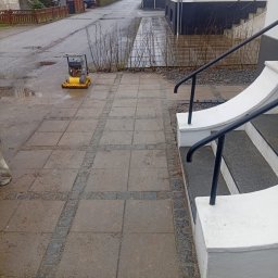 Marian Moskwa Contractor - Żółta zagęszczarka na świeżo ułożonej kostce brukowej przed domem z białymi schodami i czarną balustradą. Ulica z kałużami w tle.