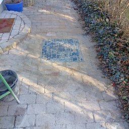 Marian Moskwa Contractor - Ścieżka z kostki brukowej w Margoninie, z widoczną studzienką kanalizacyjną i przygotowanym podłożem. Obok rosną krzewy, a w tle widać donice z roślinami.