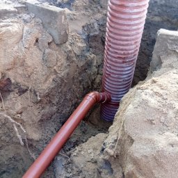 Marian Moskwa Contractor - Brązowe rury kanalizacyjne w wykopie ziemnym, pionowa rura karbowana łączy się z poziomą gładką rurą. Widoczne fragmenty betonowych obrzeży.