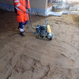 Marian Moskwa - Pracownik w pomarańczowym kombinezonie zagęszcza piasek zagęszczarką wibracyjną. Widoczne fundamenty budynku w tle. Etap przygotowania podłoża pod dalsze prace.