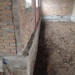 Marian Moskwa - Wykopany wąski rów wzdłuż ceglanej ściany budynku z widocznymi instalacjami wodnymi. Przygotowanie do izolacji lub drenażu fundamentów. Wnętrze budynku w trakcie prac.