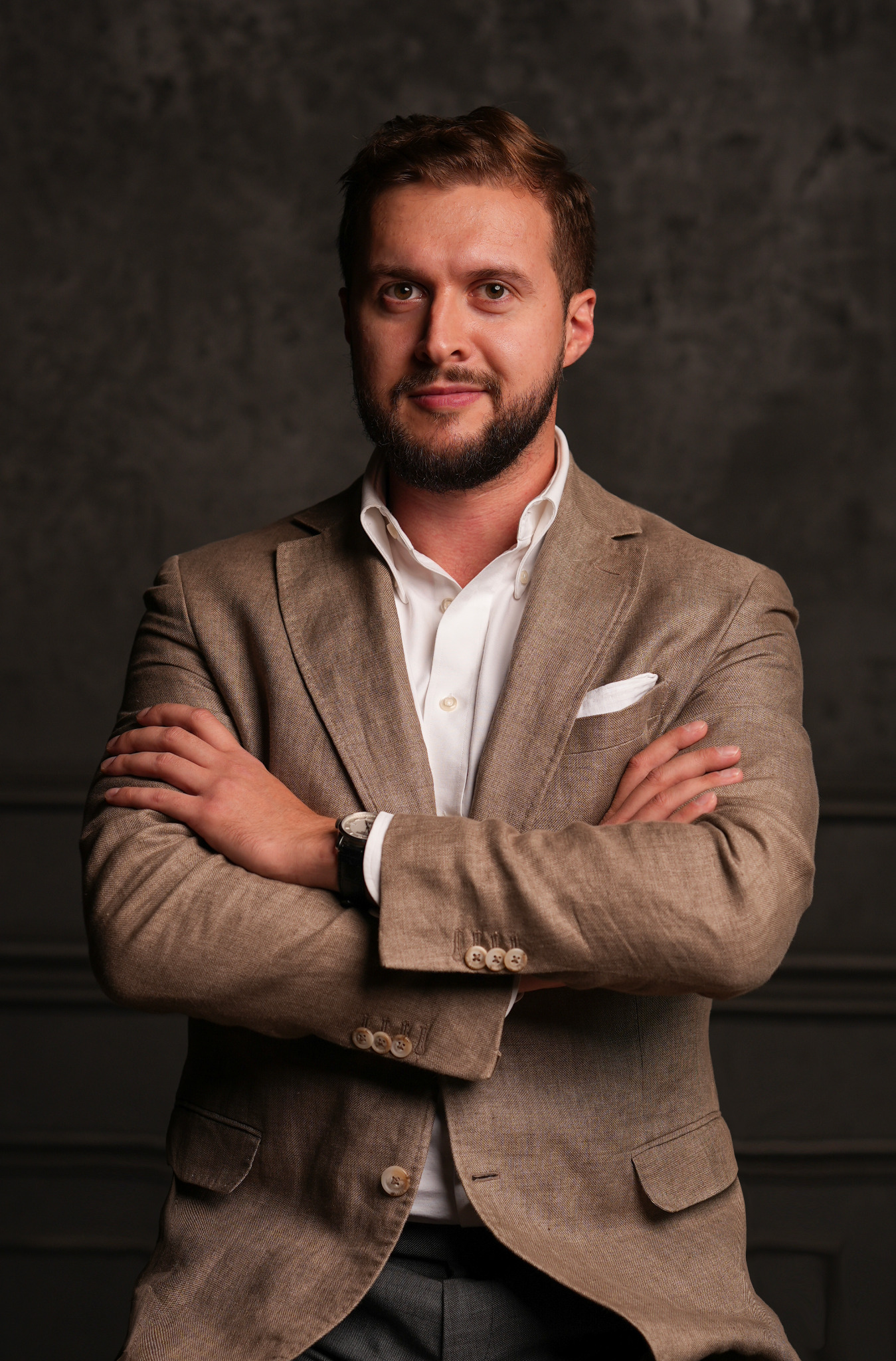 Portret mężczyzny w beżowym garniturze i białej koszuli, z zegarkiem na ręku, na tle ciemnej ściany. Skrzyżowane ręce dodają pewności.