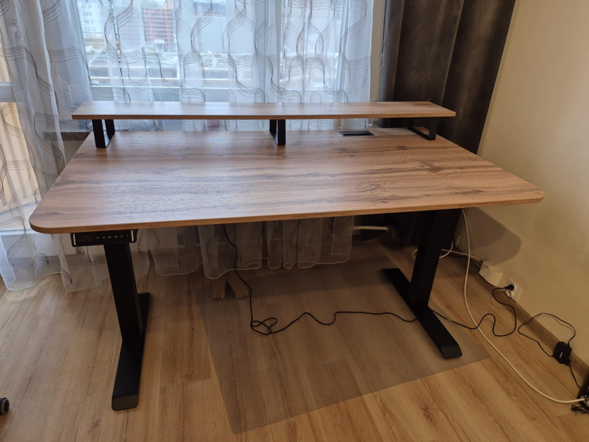 Regulowane biurko z blatem imitującym drewno i czarnymi nogami, ustawione w pokoju z zasłonami. Widoczne przewody pod biurkiem.