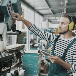Etat-Hr - Pracownik w słuchawkach obsługuje maszynę przemysłową, trzymając tablet. Ubrany w roboczy kombinezon i bluzę w paski, w tle hala produkcyjna z maszynami.