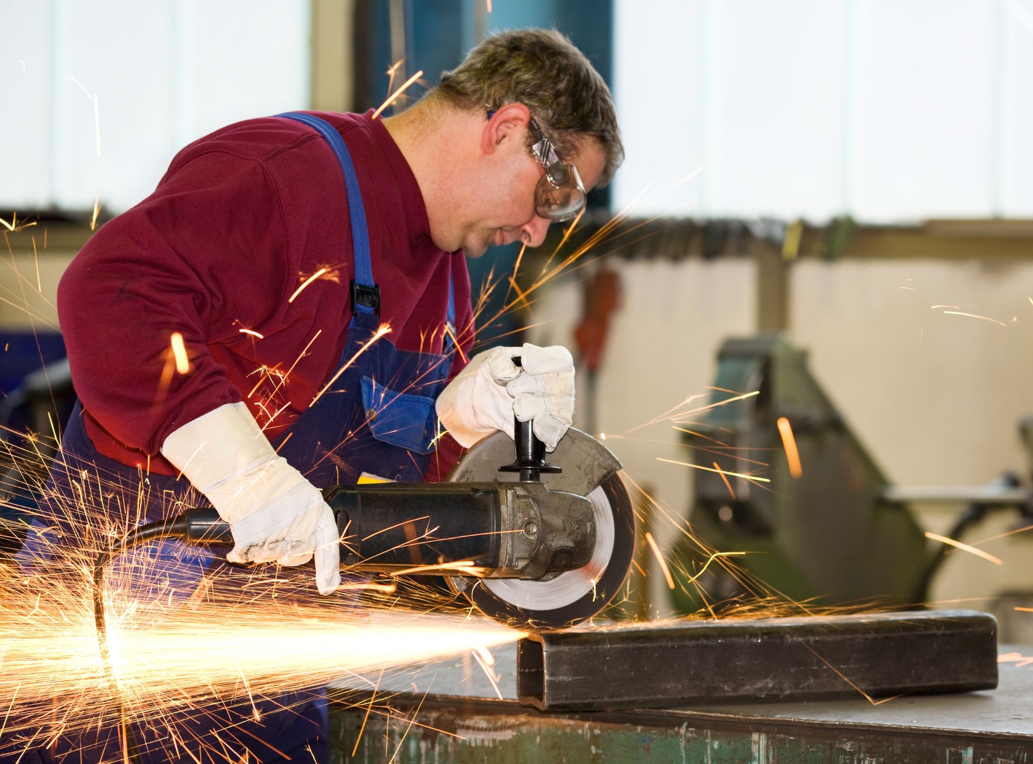 Pracownik w okularach ochronnych szlifuje metalową konstrukcję szlifierką kątową, iskry rozbryzgują się na boki. Praca z metalem, bezpieczeństwo, odzież robocza.