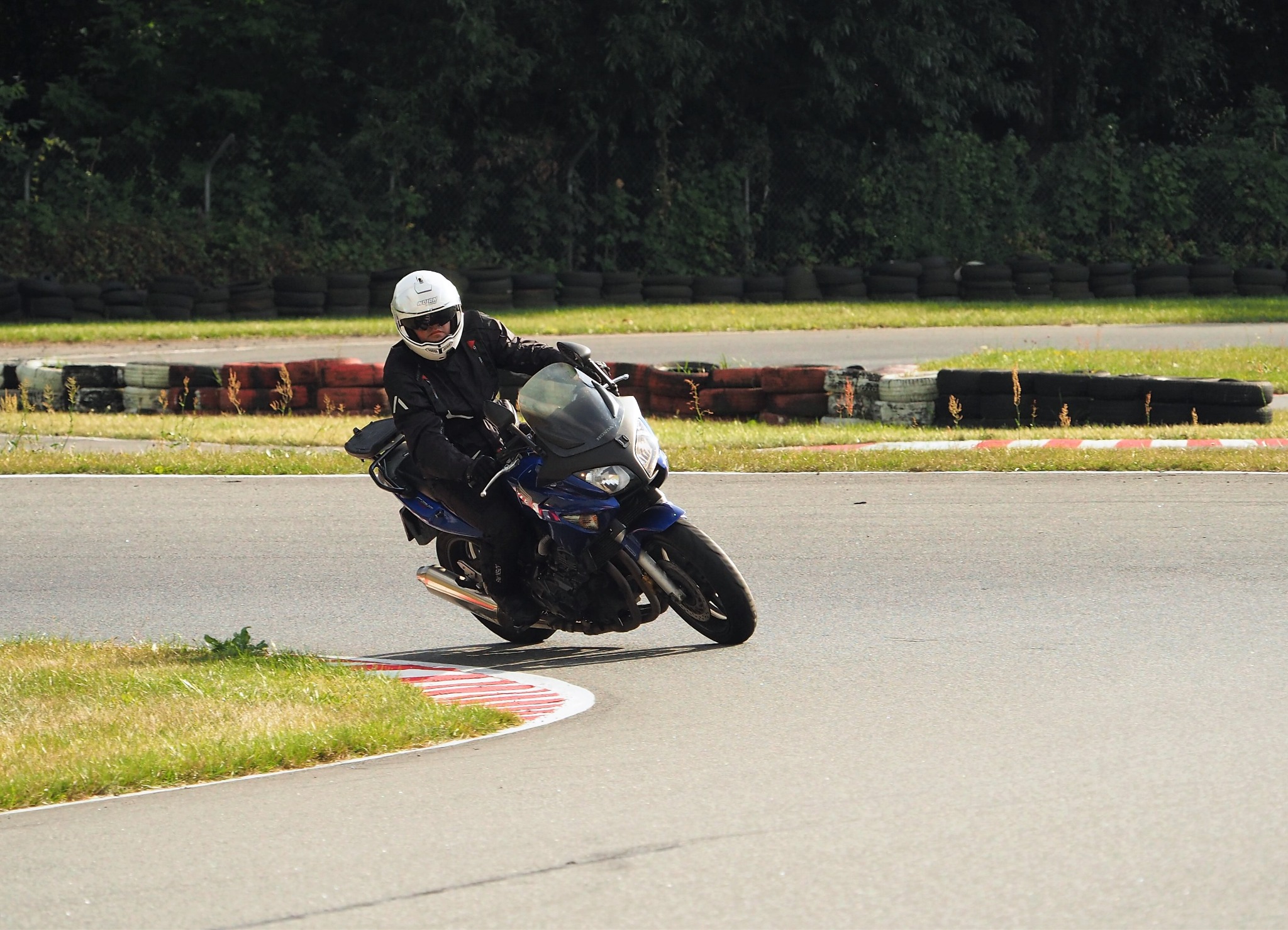 Motocyklista w białym kasku pokonuje zakręt na torze, w tle opony i zieleń. Dynamiczne ujęcie z bliska, ukazujące pochylenie motocykla i skupienie kierowcy.