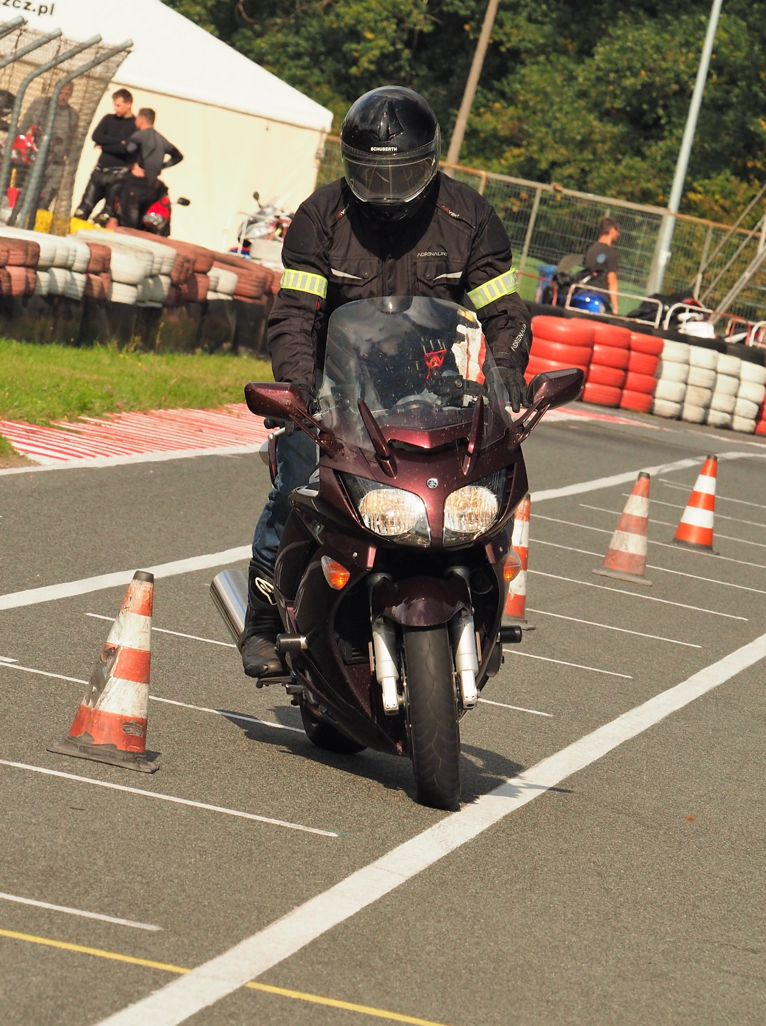Motocyklista w pełnym stroju na torze, pokonuje slalom między pachołkami. W tle opony i osoby obserwujące. Ujęcie z przodu, w słoneczny dzień.
