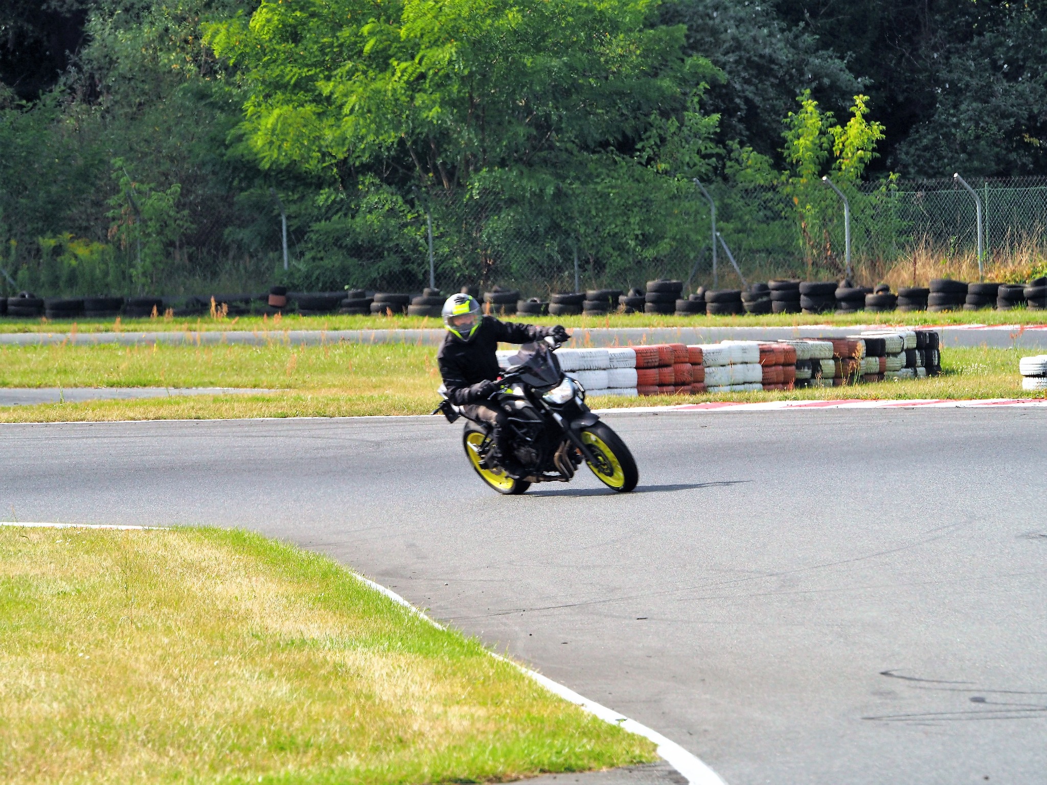Motocyklista w czarnym kombinezonie i żółtym kasku pokonuje zakręt na torze wyścigowym, otoczonym oponami i zielenią. Dynamiczne ujęcie z bliska.