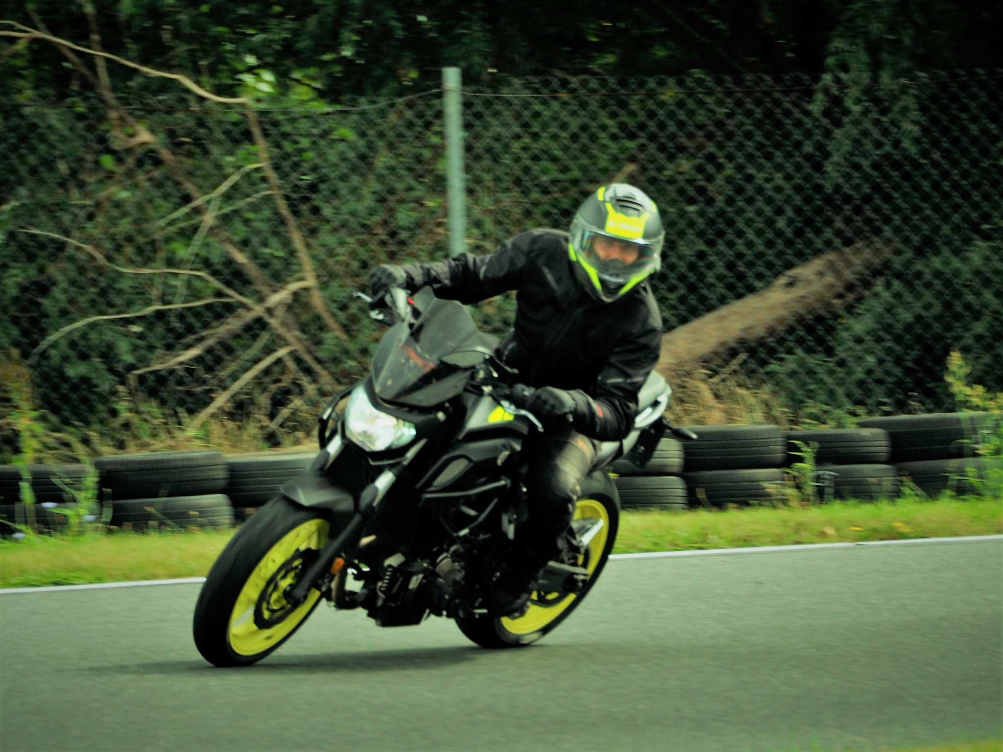 Motocyklista w kasku i kombinezonie na czarnym motocyklu z żółtymi felgami, pokonujący zakręt na torze. W tle opony i zieleń.