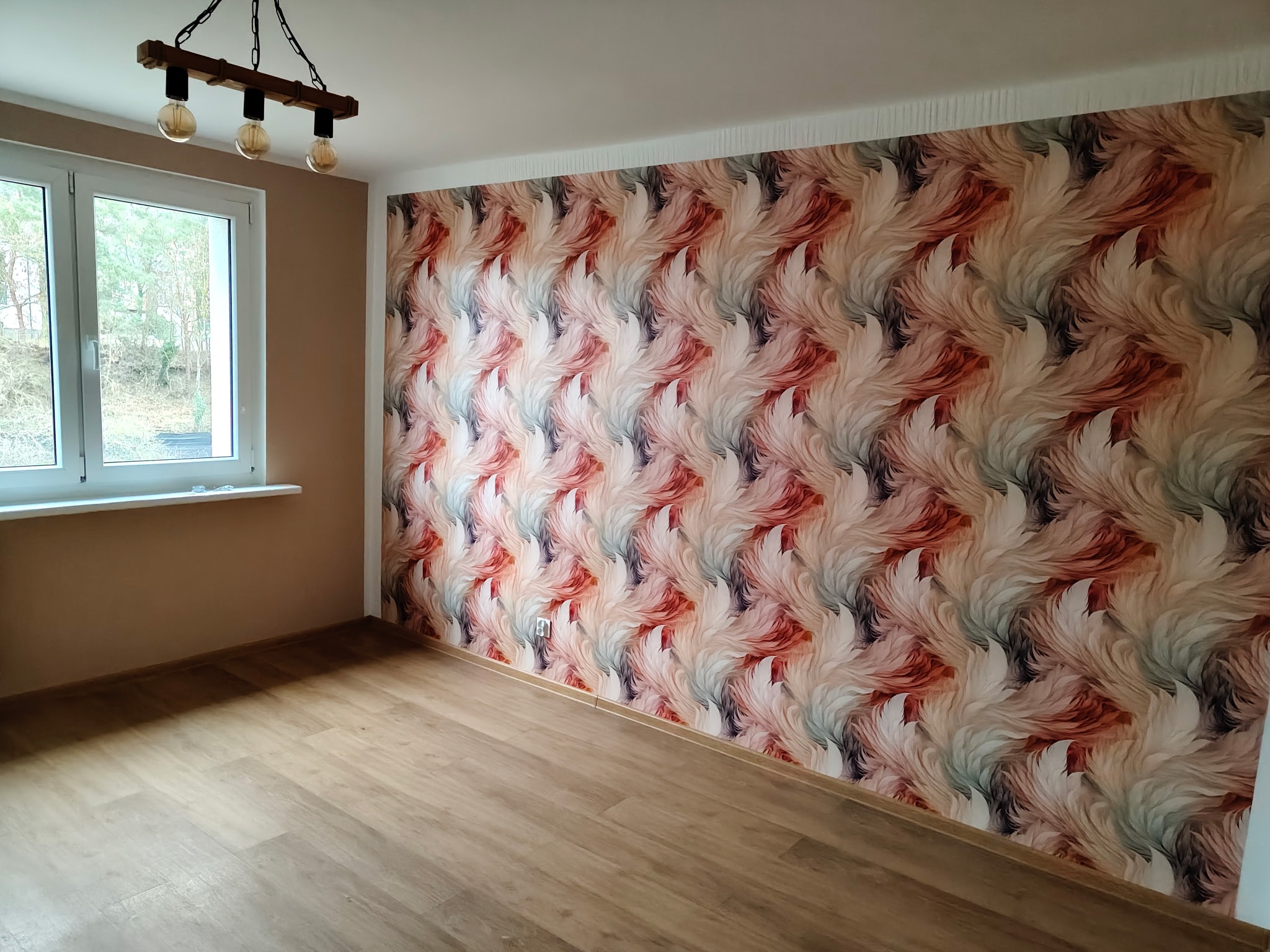 Pokój po remoncie: wzorzysta tapeta w odcieniach bieli, różu i brązu na ścianie, drewniana podłoga, okno z widokiem na zieleń i lampa z trzema żarówkami.