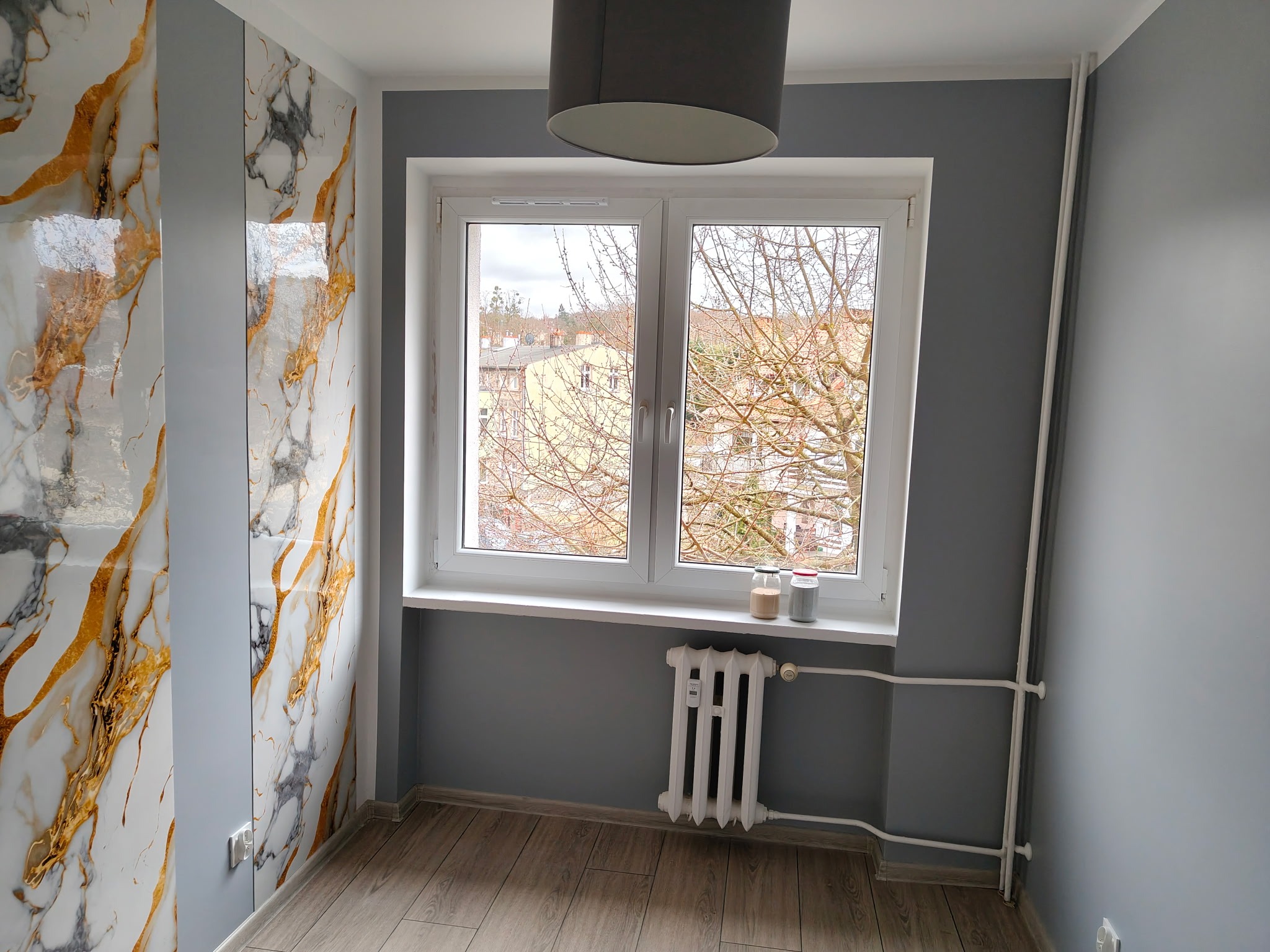 Pokój po remoncie: szare ściany, panele podłogowe, białe okno z widokiem na drzewa, grzejnik i ściana z efektem marmuru w odcieniach bieli i złota.