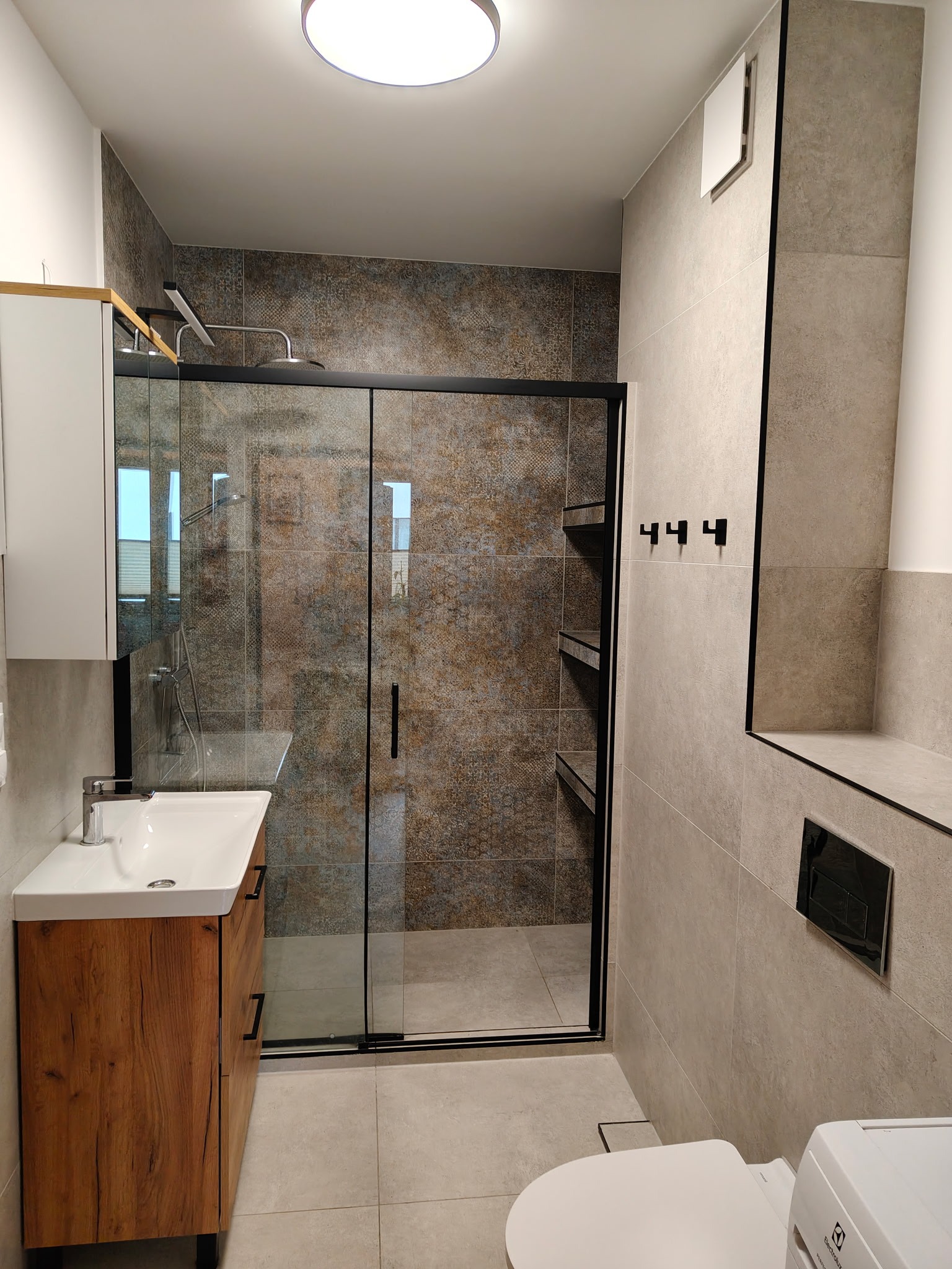 Nowoczesna łazienka po remoncie: kabina prysznicowa ze szklanymi drzwiami, umywalka z szafką z drewnopodobnym frontem, beżowe płytki i czarne akcenty.