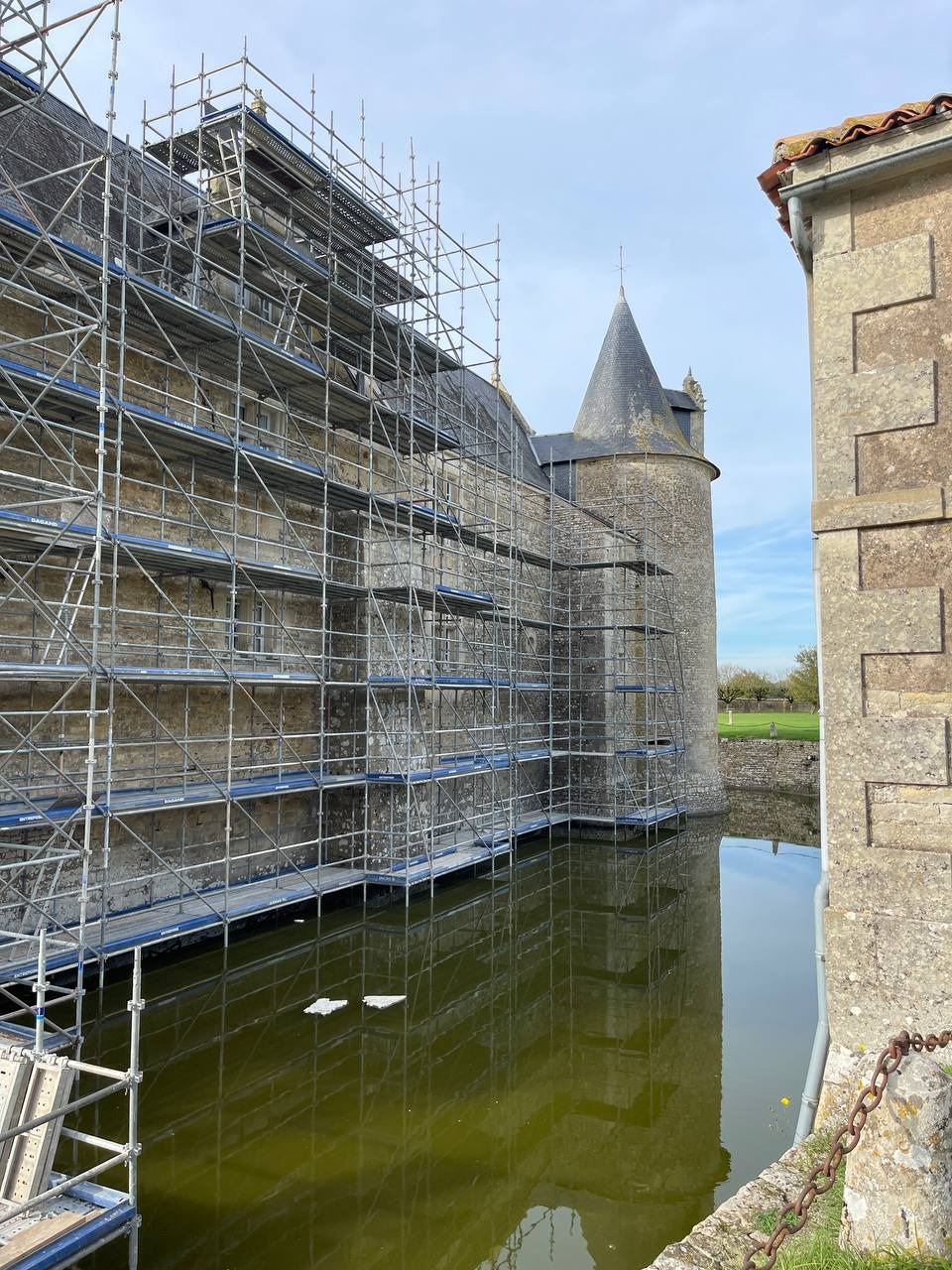 Zamek otoczony rusztowaniami nad fosą, przygotowywany do prac. Kamienna elewacja w trakcie renowacji, odbicie w wodzie. Błękitne niebo w tle.