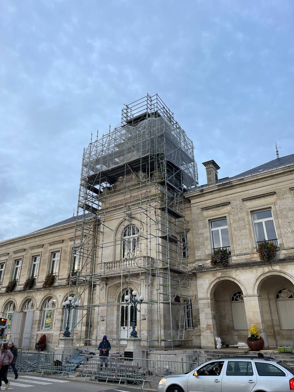 Budynek miejski w trakcie prac elewacyjnych, otoczony rusztowaniami. Widoczne detale architektoniczne i elementy zabezpieczające. Pochmurne niebo w tle.