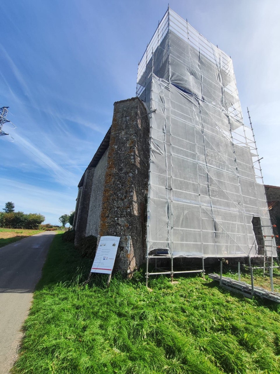 Kamienna wieża kościoła w trakcie prac. Część budynku osłonięta rusztowaniami i siatką ochronną. Tablica informacyjna przy drodze. Błękitne niebo w tle.