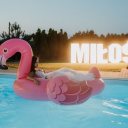 FOKUS GROUP Michał Margas - Panna młoda w sukni na różowym flamingowym materacu w basenie, w tle podświetlony napis 'Miłość' i mężczyzna w fotelu. Romantyczna sceneria ślubna.