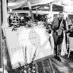 FOKUS GROUP Michał Margas - Czarno-białe zdjęcie saksofonisty w garniturze grającego na saksofonie na weselu, z tablicą powitalną z imionami pary młodej na pierwszym planie.