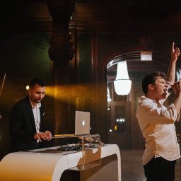 FOKUS GROUP Michał Margas - Zespół muzyczny na scenie: wokalista z mikrofonem, skrzypek i DJ z laptopem. W tle dekoracyjne, ciemne ściany i oświetlenie sceniczne.