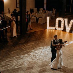 FOKUS GROUP Michał Margas - Para młoda tańczy na parkiecie w sali weselnej, oświetlona dekoracyjnym napisem LOVE. Goście obserwują, w tle stoły i kolumny.