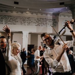 FOKUS GROUP Michał Margas - Uśmiechnięty skrzypek grający na elektrycznych skrzypcach z fajerwerkami na weselu, w tle tańczący goście i dekoracje sali.